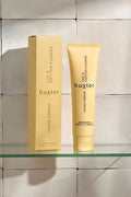 A yellow tube of Huxter hand cream in the scent "Lily & Cotton Flower" stands beside its matching rectangular box on a clear glass shelf, against a soft-tiled beige wall with natural light casting gentle shadows.