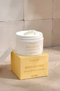 An open white jar of Huxter body butter in the scent "Lily & Cotton Flower" rests on top of its soft yellow box, revealing a smooth, whipped cream texture inside. The setup sits on a warm-toned surface against a tiled beige wall, bathed in natural light and soft shadows.