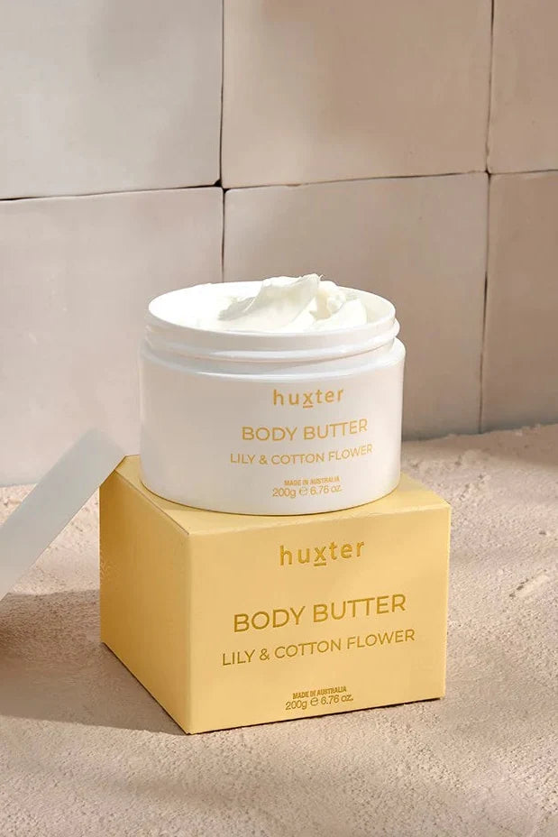 An open white jar of Huxter body butter in the scent "Lily & Cotton Flower" rests on top of its soft yellow box, revealing a smooth, whipped cream texture inside. The setup sits on a warm-toned surface against a tiled beige wall, bathed in natural light and soft shadows.