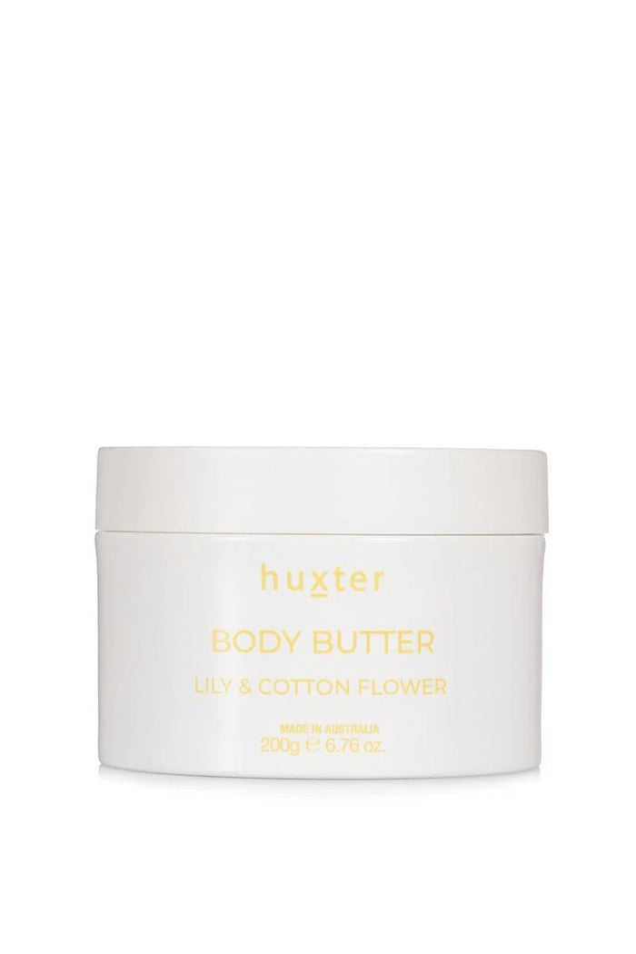 An open white jar of Huxter body butter in the scent "Lily & Cotton Flower" rests on top of its soft yellow box, revealing a smooth, whipped cream texture inside. The setup sits on a warm-toned surface against a tiled beige wall, bathed in natural light and soft shadows.