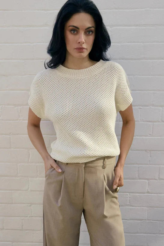 Woman wearing a beige knitted top and beige pants against a white brick wall.