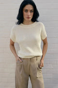 Woman wearing a beige knitted top and beige pants against a white brick wall.