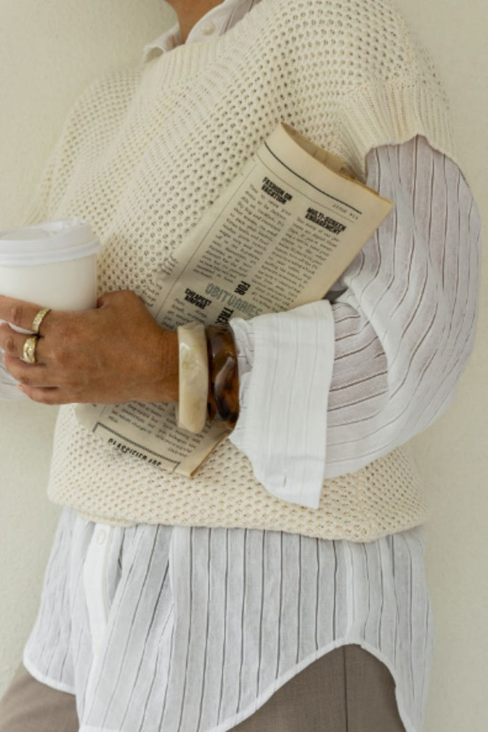 Person wearing a beige sweater over a white shirt, holding a newspaper and a cup.