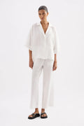 A model wearing a white short-sleeve button-up shirt and matching white wide-leg pants, paired with black slide sandals, against a plain white studio background.