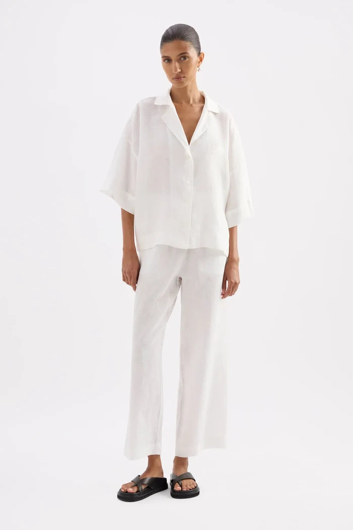A model wearing a white short-sleeve button-up shirt and matching white wide-leg pants, paired with black slide sandals, against a plain white studio background.