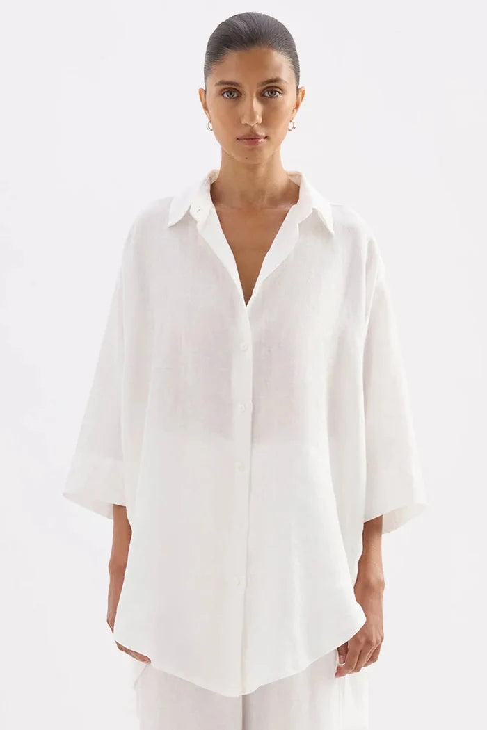 A model stands against a plain white studio background wearing a loose white button-up shirt with three-quarter sleeves and matching wide-leg white pants, paired with black sandals.