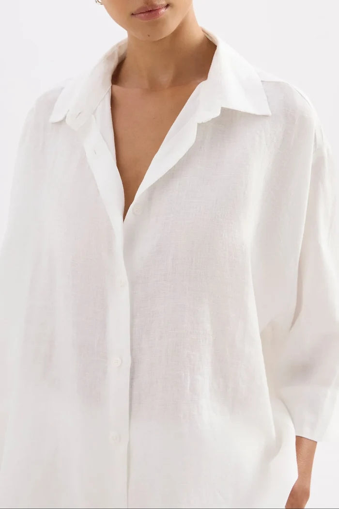 A close-up of a model wearing an oversized white linen button-up shirt with a pointed collar and three-quarter sleeves.