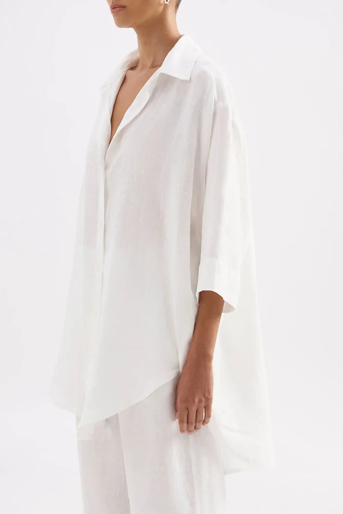 A model wearing a loose white linen button-up shirt with three-quarter sleeves and white trousers, shown from the side against a plain white background.