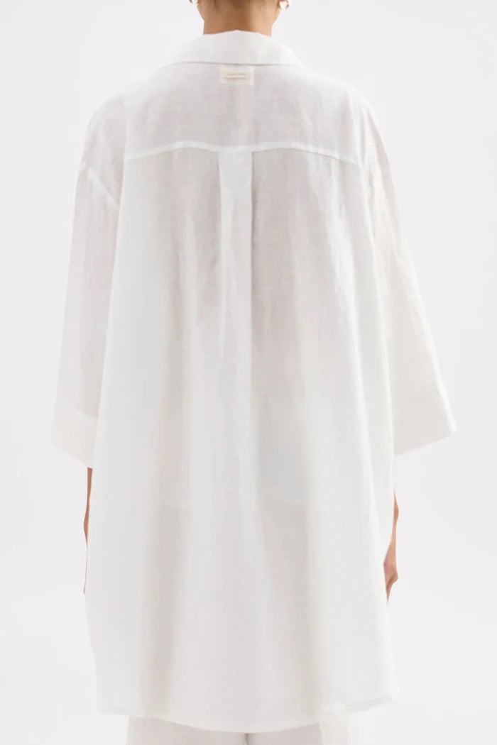 A person shown from the back wearing a loose white linen shirt with three-quarter sleeves and a wide collar, photographed against a clean white studio background.
