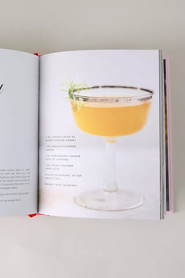 An open recipe book showing a cocktail recipe page with a photograph of a yellow-orange margarita in a coupe glass with a salted rim and a sprig of rosemary garnish.