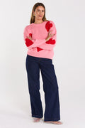 Woman wearing a pink sweater with red heart patterns and dark blue pants on a white background