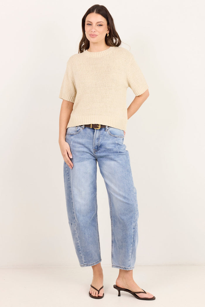 A woman modeling a beige short-sleeve knit top and light blue cropped jeans with black sandals, standing against a plain off-white studio background.