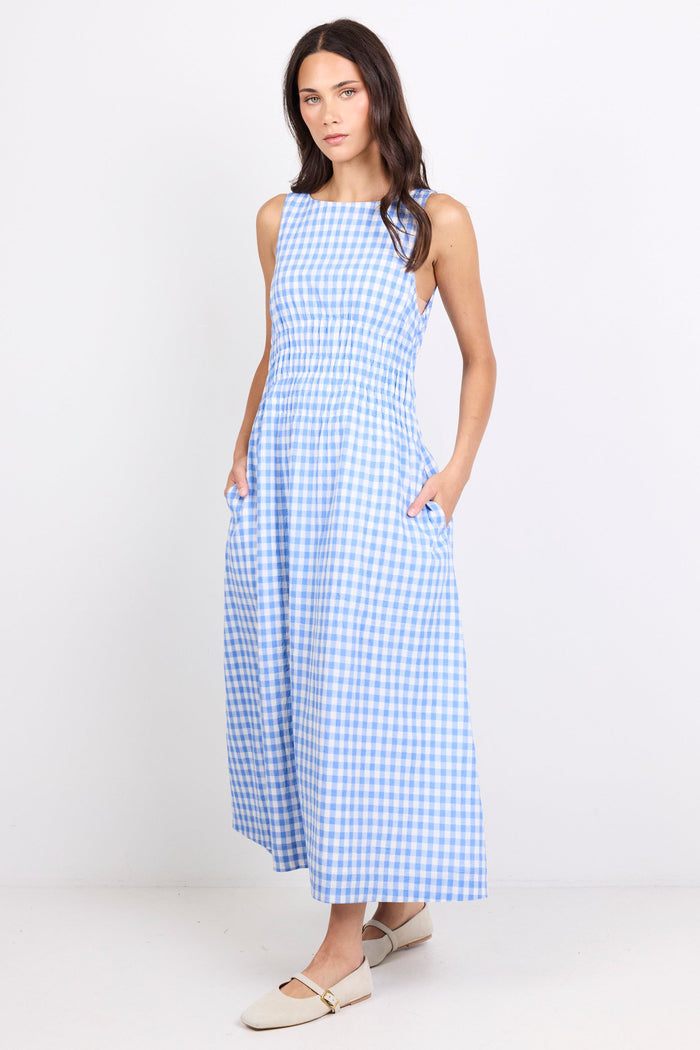 A woman modeling a sleeveless, high-neck, midi-length dress with a blue and white gingham check pattern, standing against a plain white studio background.