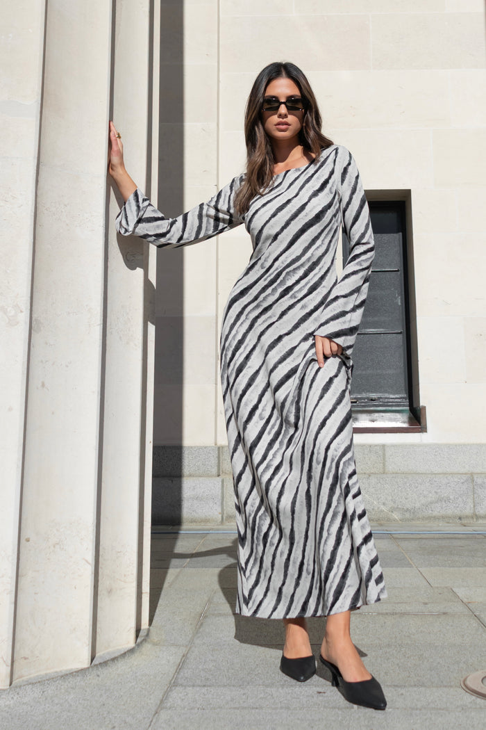 Woman wearing a full-length bias-cut dress with long sleeves and a diagonal charcoal stripe print, styled with black pointed heels.