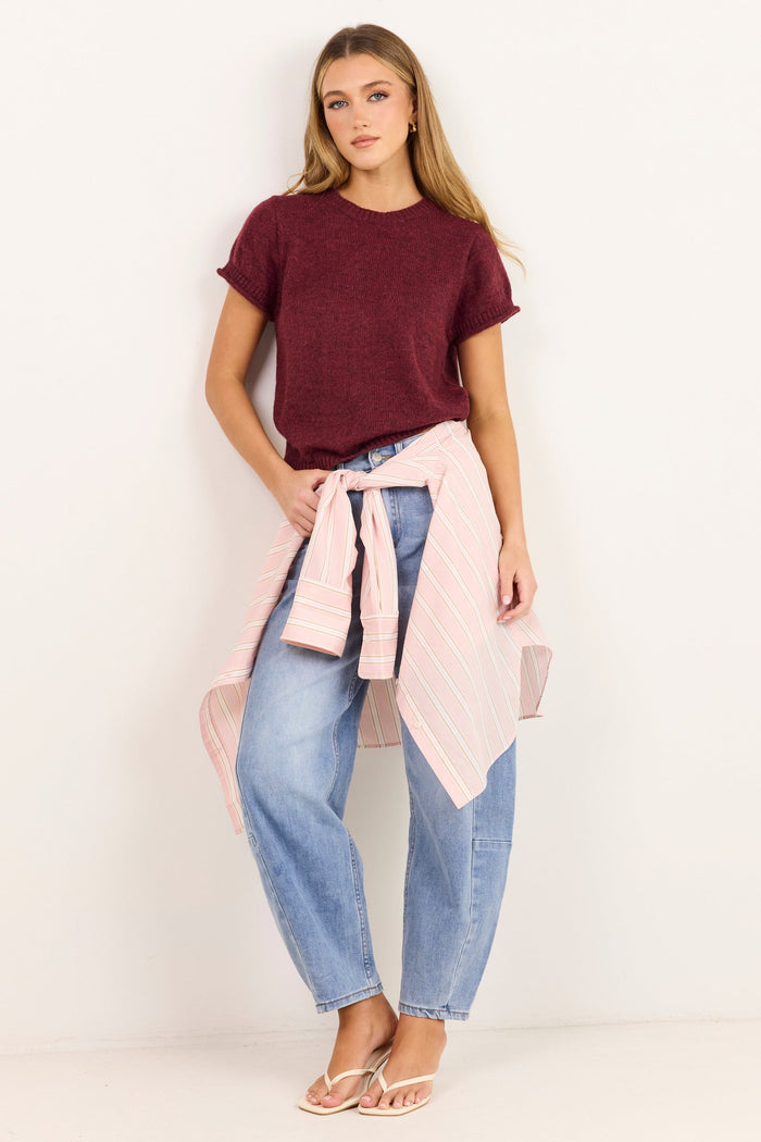 A woman modeling a dark burgundy short-sleeve t-shirt, light-wash blue jeans, and a light pink striped shirt tied around her waist, standing against a plain white studio background.