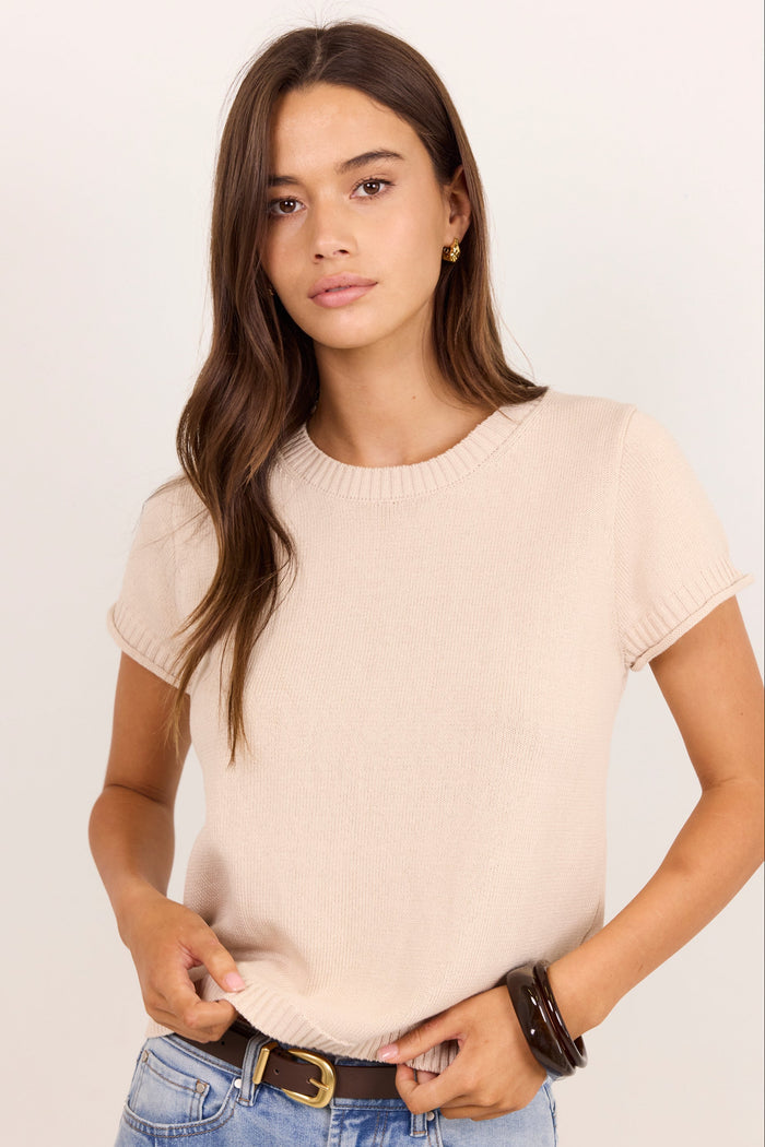 A woman modeling a short-sleeve crewneck knit top in a light beige color, paired with blue jeans and a brown belt, against a plain light background.