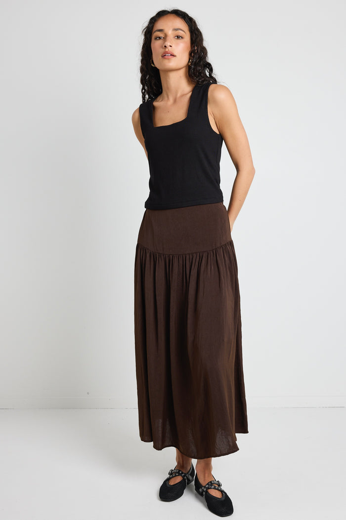 A woman wearing a flowing espresso-coloured hammered satin skirt with soft gathers, styled with a black top and textured flats.