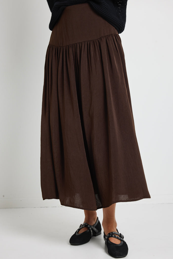 A woman wearing a flowing espresso-coloured hammered satin skirt with soft gathers, styled with a black top and textured flats.
