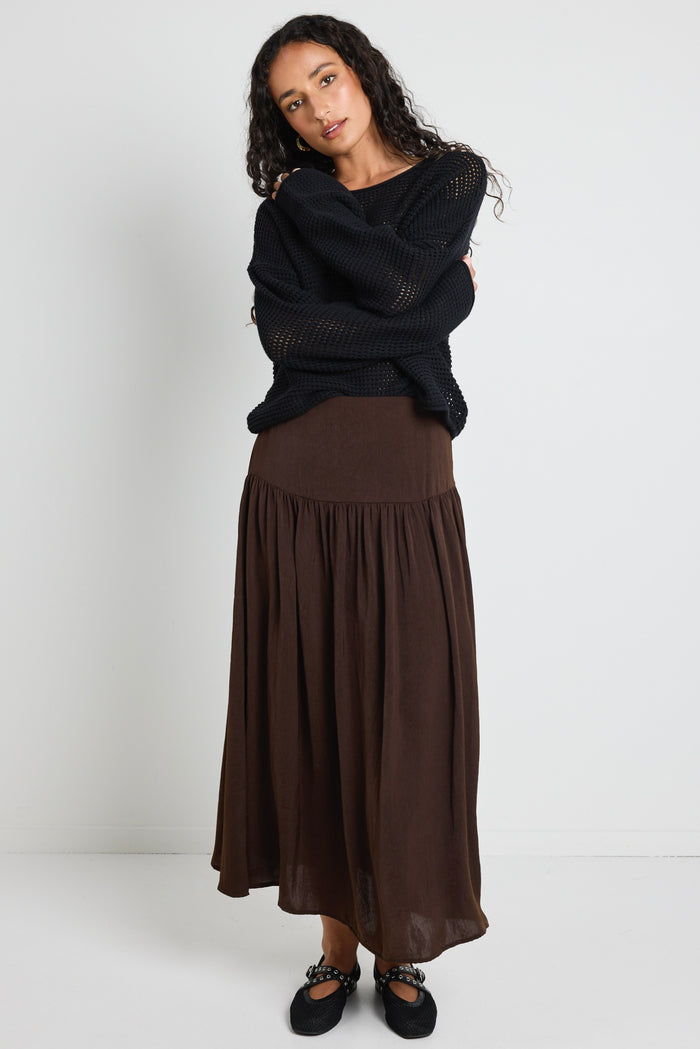 A woman wearing a flowing espresso-coloured hammered satin skirt with soft gathers, styled with a black top and textured flats.