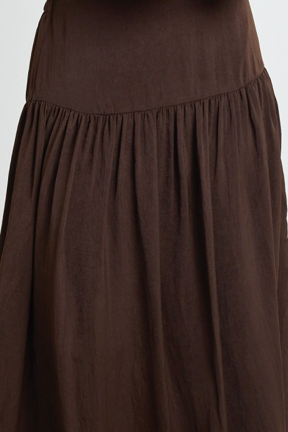 A woman wearing a flowing espresso-coloured hammered satin skirt with soft gathers, styled with a black top and textured flats.