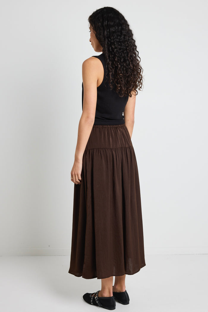 A woman wearing a flowing espresso-coloured hammered satin skirt with soft gathers, styled with a black top and textured flats.