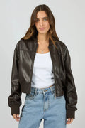 Woman wearing a brown leather jacket, white shirt, and blue jeans on a gray background