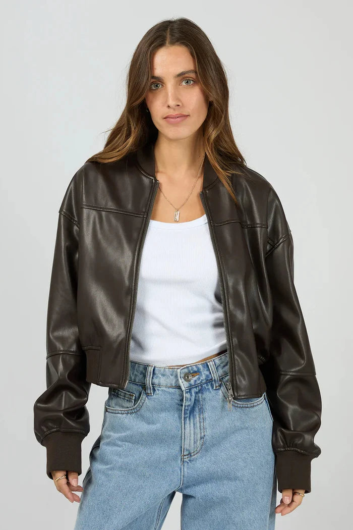 Woman wearing a brown leather jacket, white shirt, and blue jeans on a gray background