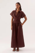 A woman modeling a dark brown midi dress with short flutter sleeves, a collared neckline, a gathered waist, and a flowing skirt, set against a plain light gray studio background.