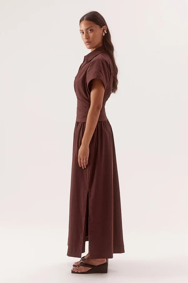 A woman modeling a dark brown short-sleeve collared maxi dress with a fitted waist and side slit, paired with dark wedge sandals, against a plain light background.
