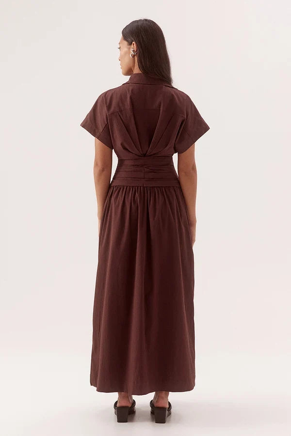 A woman viewed from the back wearing a dark brown short-sleeve midi dress with a gathered waist and pleated back detail, paired with black heels, against a plain light background.