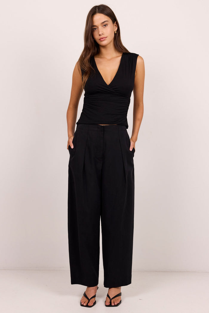 A woman modeling a black sleeveless wrap-style top and matching black wide-leg trousers, standing against a plain light studio background.