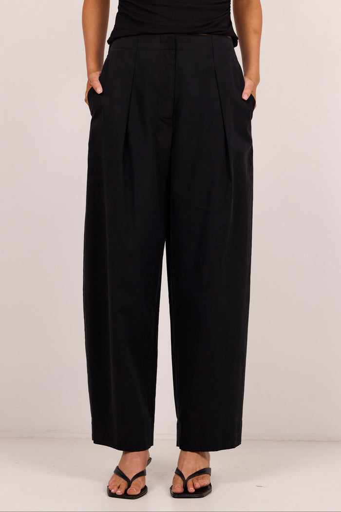 A model wearing black wide-leg cropped trousers with side pockets, paired with a black top and black sandals, standing against a plain off-white studio background.
