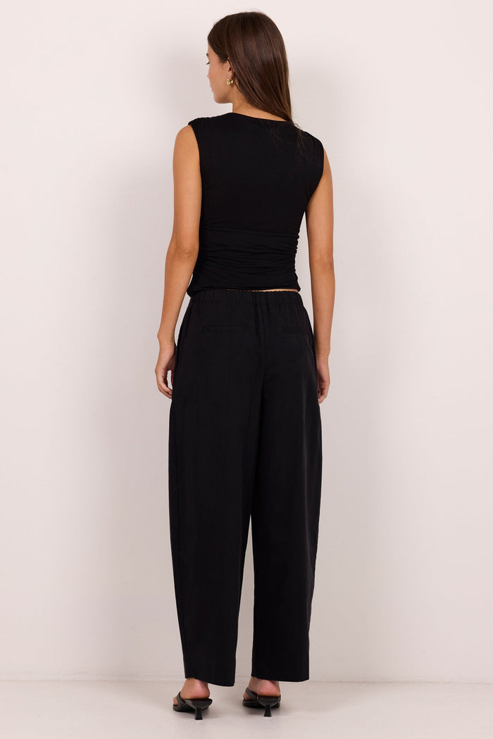 A woman viewed from the back wearing a black sleeveless top and black wide-leg pants, standing against a plain off-white studio wall.