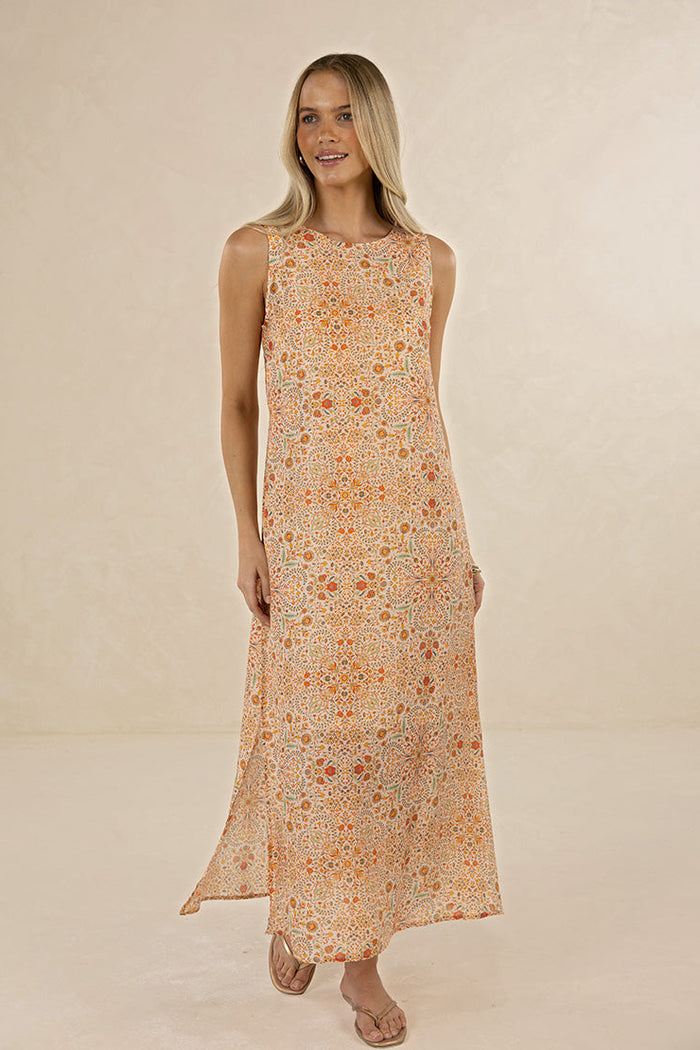 A woman modeling a sleeveless, floor-length maxi dress with an intricate orange, yellow, and beige floral mandala pattern against a warm beige studio backdrop.