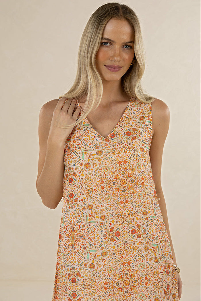 A blonde woman wearing a sleeveless V-neck dress with an intricate orange, yellow, and beige floral pattern, posed against a solid beige background.