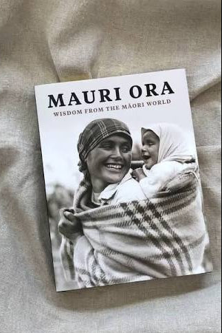 A book titled "MAURI ORA: WISDOM FROM THE MÁORI WORLD" lying on a textured beige fabric background. The cover features a black-and-white photograph of a smiling Máori woman carrying a child wrapped in a plaid blanket.