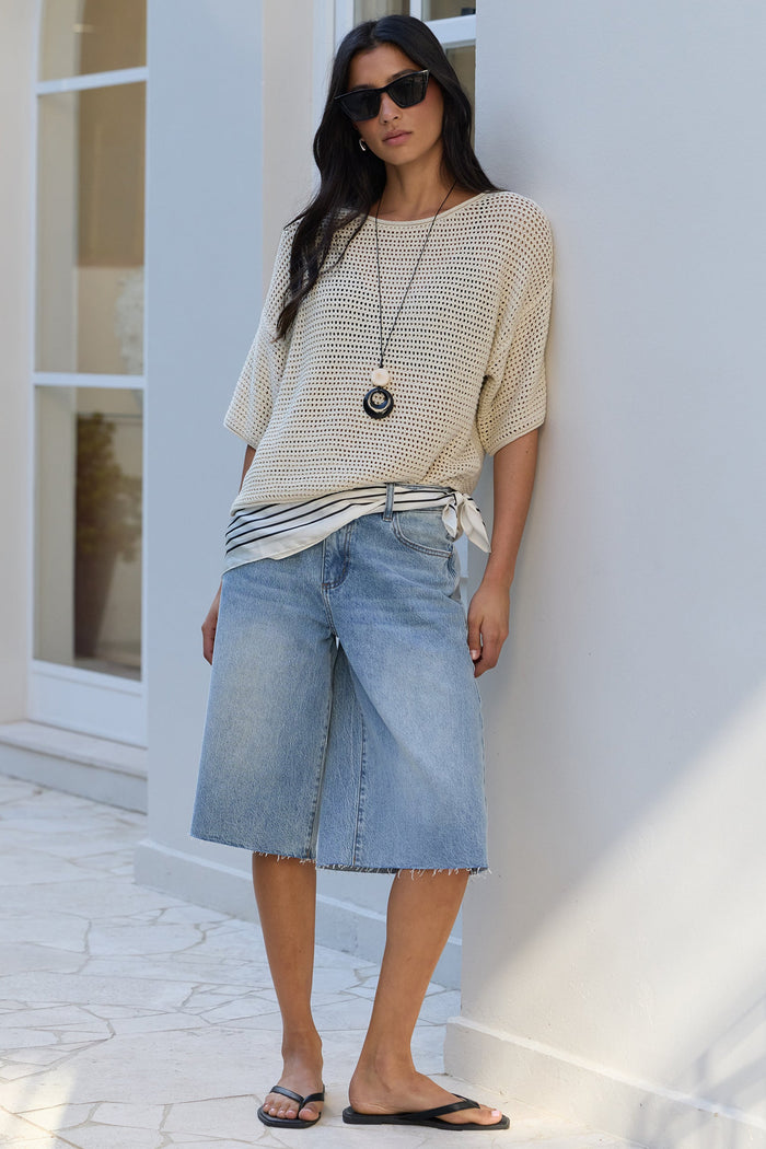 A woman stands outdoors against a white building wall wearing a cream knit top, light-wash denim shorts, black flip-flops, and sunglasses.