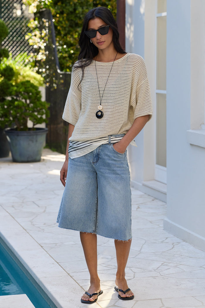 A woman stands by a pool wearing a cream short-sleeve knit top, light-wash denim cutoff shorts, sunglasses, and flip-flops in a bright outdoor patio setting.