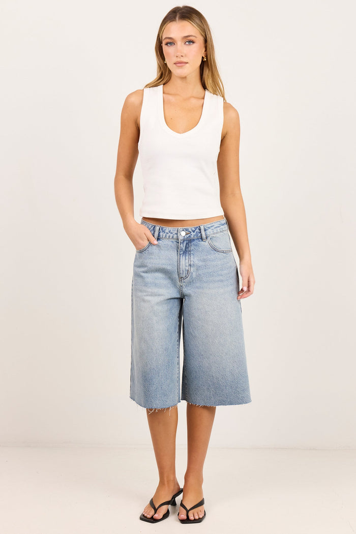 A woman modeling a white ribbed tank top and light-wash blue cropped denim shorts against a plain off-white studio background.