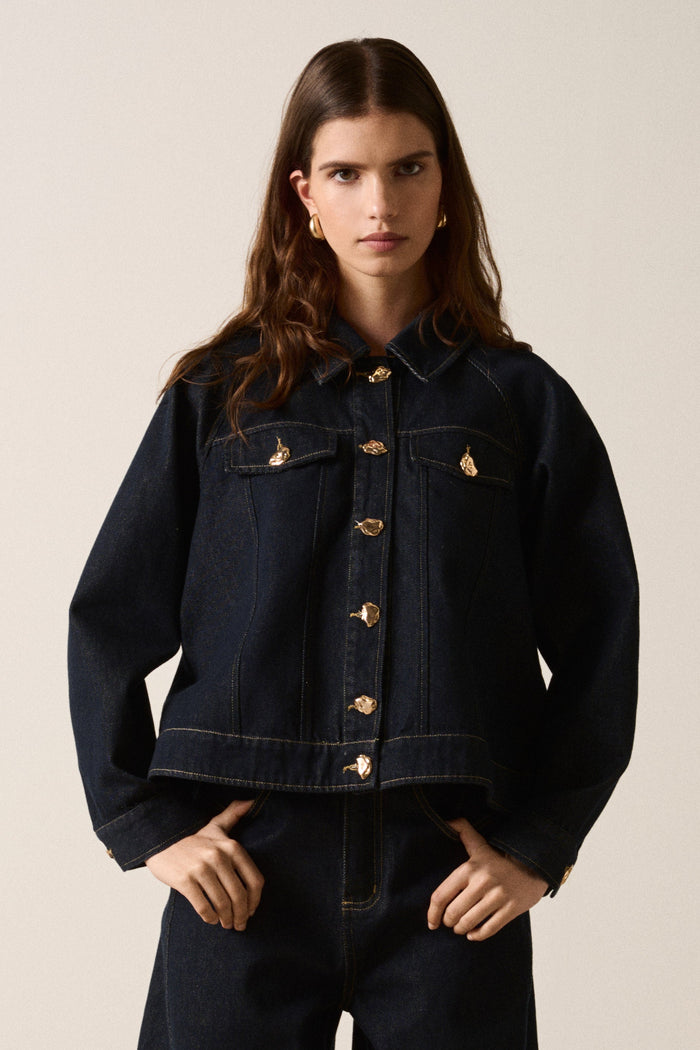 Person wearing a dark denim jacket and pants on a plain background
