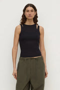 A woman modeling a black sleeveless knit top with cutout details at the shoulders and sides, paired with olive green pants, against a plain light gray studio background.