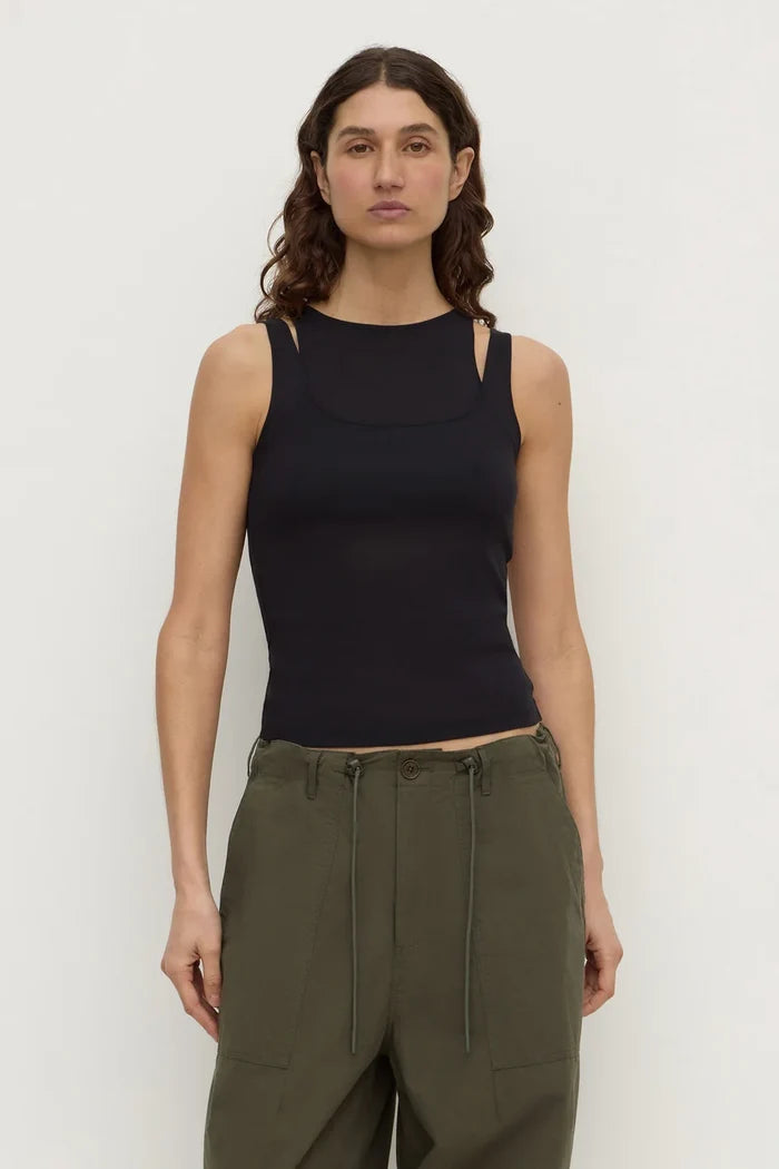 A woman modeling a black sleeveless knit top with cutout details at the shoulders and sides, paired with olive green pants, against a plain light gray studio background.