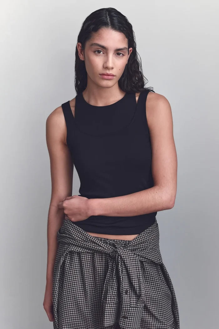 A woman modeling a black sleeveless crop top with side cutouts and a high neckline, paired with a black and white gingham shirt tied around her waist, against a light gray studio background.