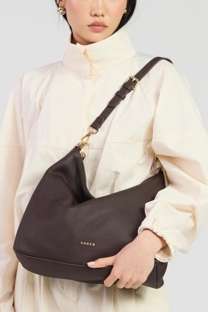 Person holding a dark brown shoulder bag with a visible brand logo against a neutral background