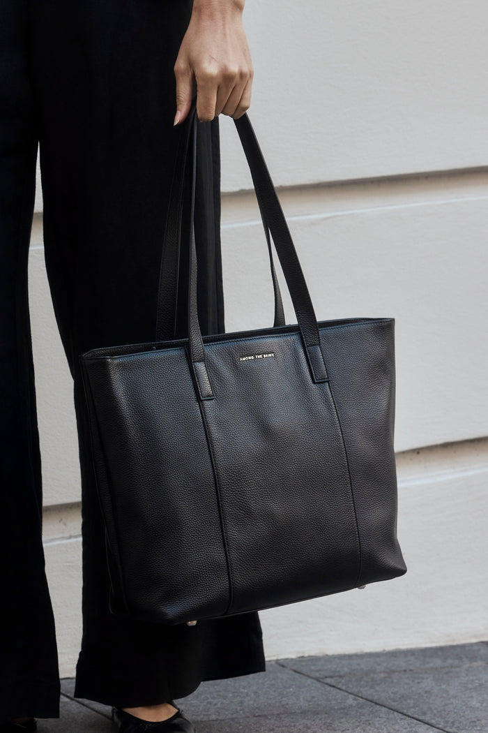 A person wearing black wide-leg trousers and a blue striped shirt holds a large black leather tote bag while standing outdoors against a light beige wall.