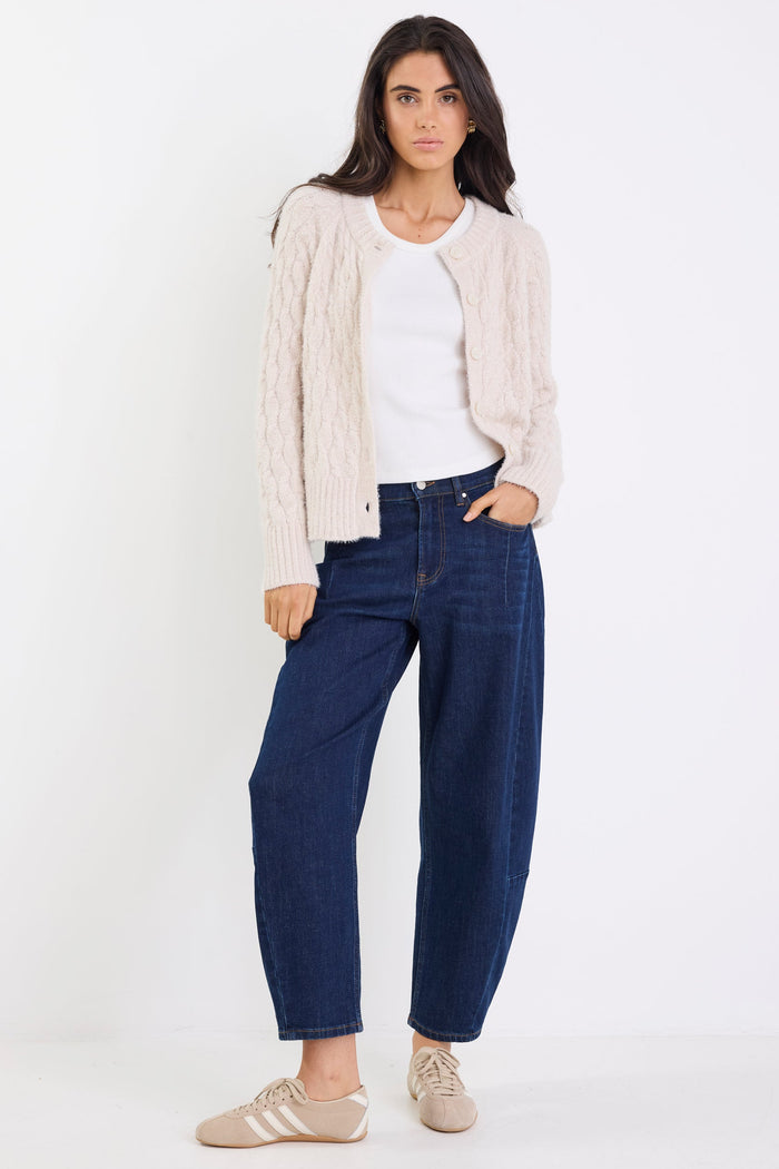 Woman wearing a beige cardigan, white shirt, and blue jeans on a white background