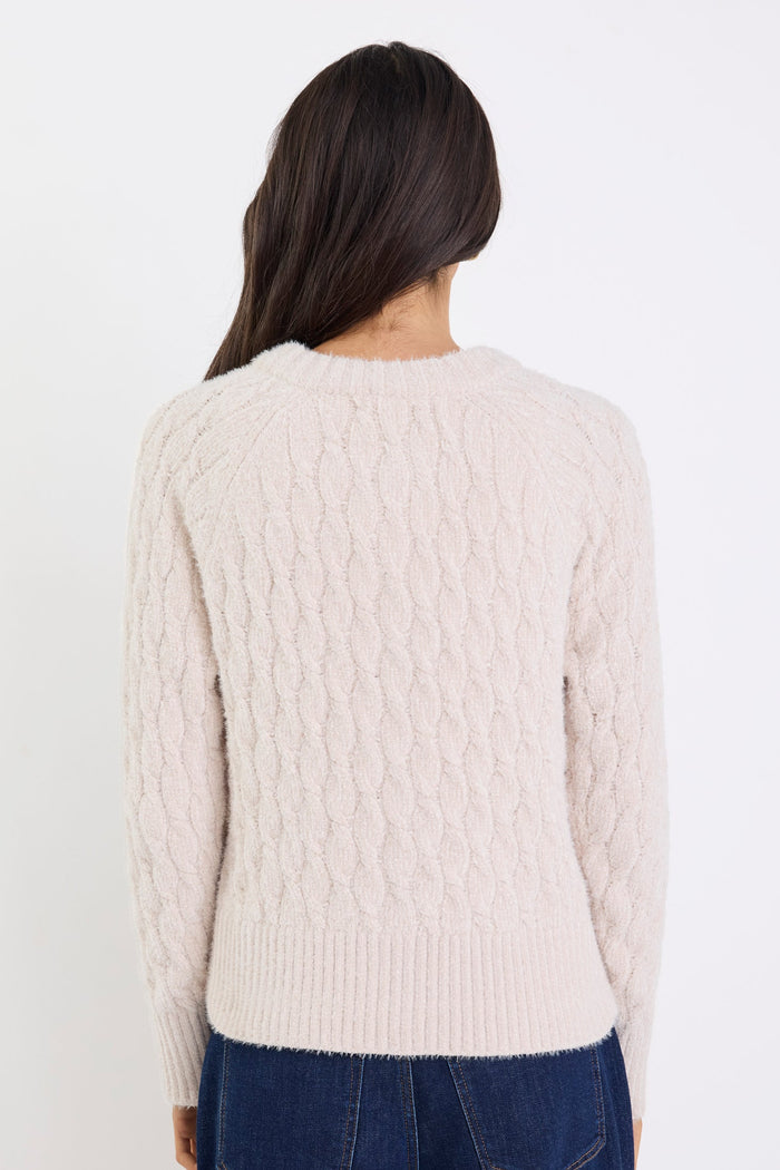 Person wearing a cream cable knit sweater and dark blue jeans on a white background