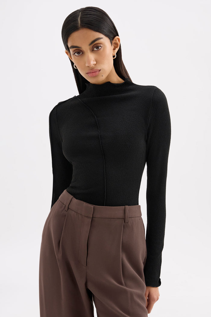 A woman modeling a black long-sleeve mock neck top paired with brown trousers, shot against a plain light gray studio background.