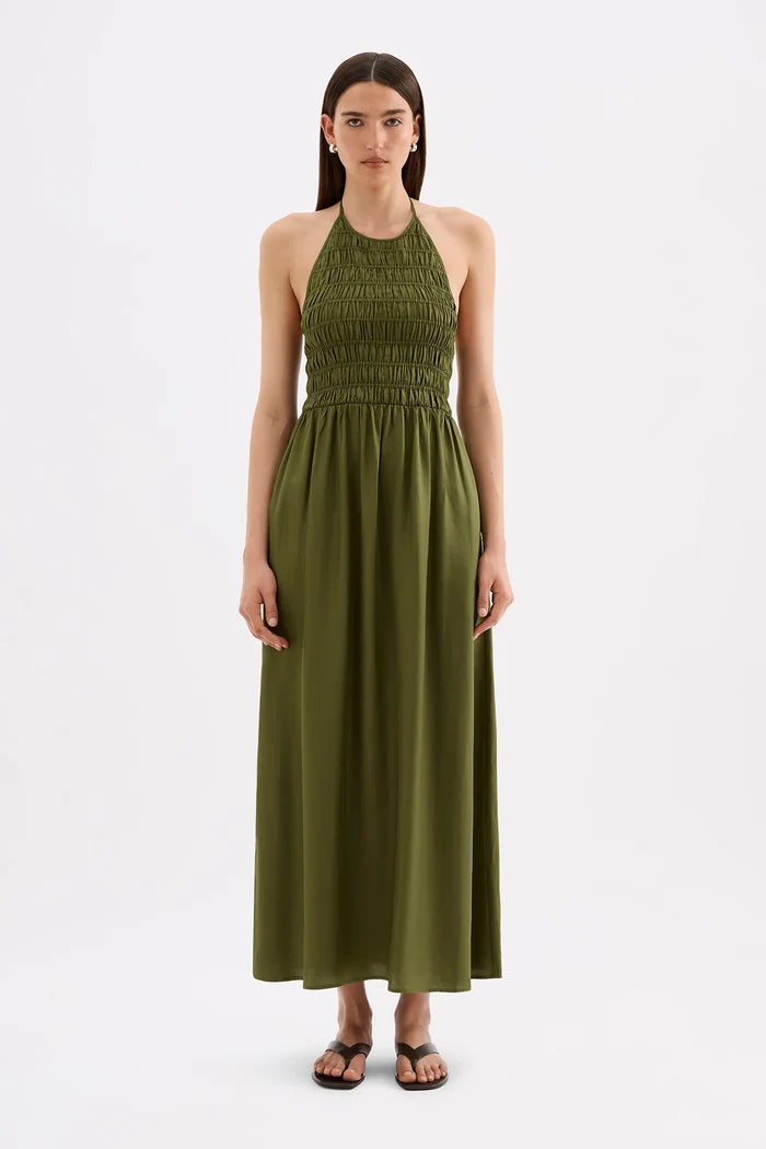 A woman modeling a sleeveless olive green halter maxi dress with a smocked bodice, standing against a plain white studio background.