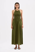 A woman modeling a sleeveless olive green halter maxi dress with a smocked bodice, standing against a plain white studio background.